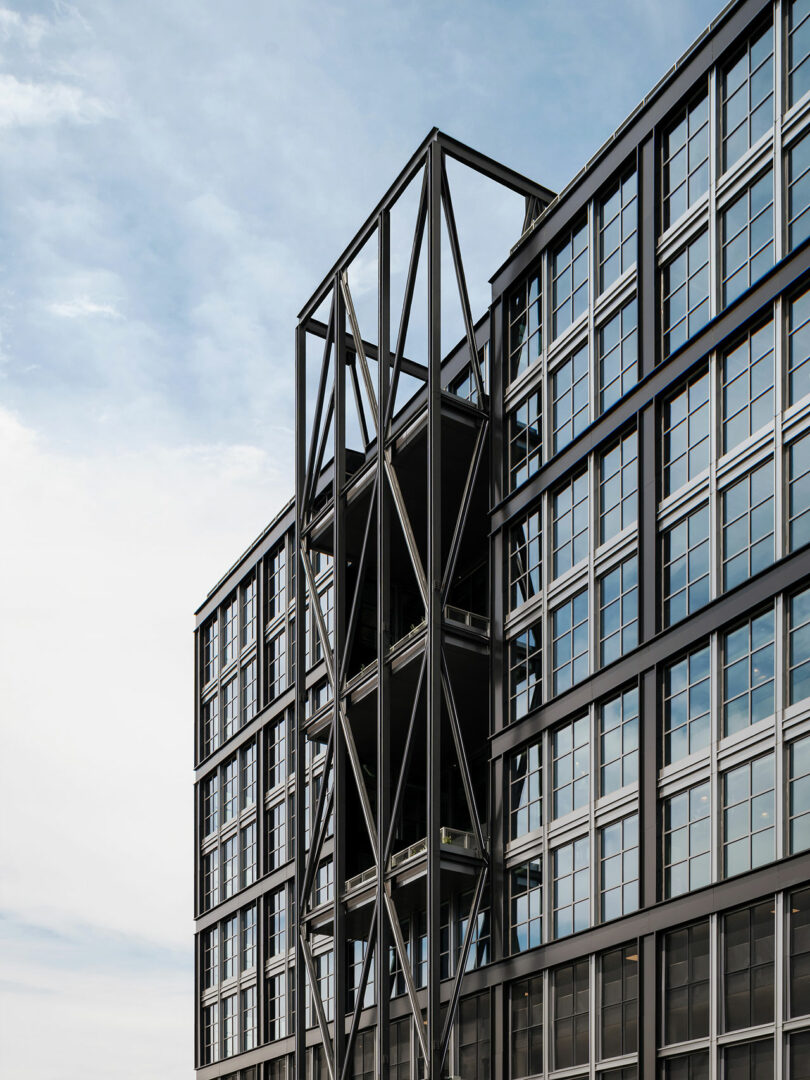 Modern glass and steel building by Morris Adjmi Architects, featuring prominent diagonal external structural supports, set against a partly cloudy sky.