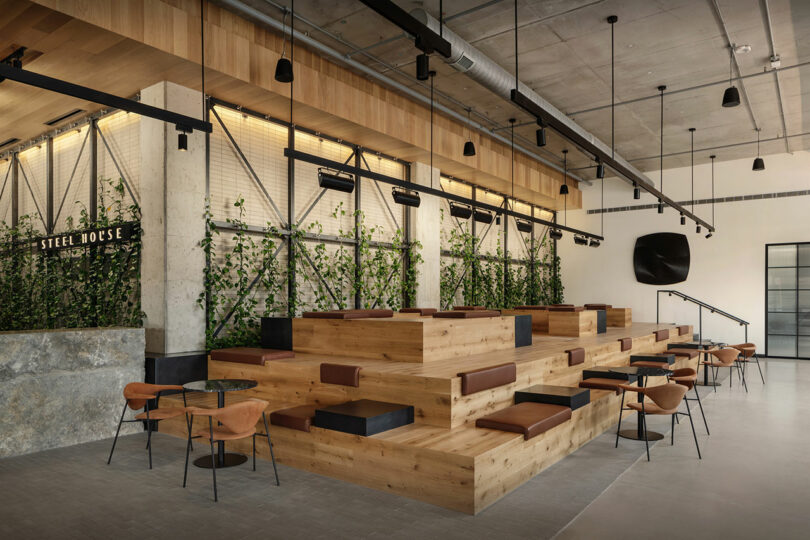 Modern café interior by Morris Adjmi Architects with tiered wooden seating, brown chairs, small round tables, greenery along the wall, and industrial ceiling details.