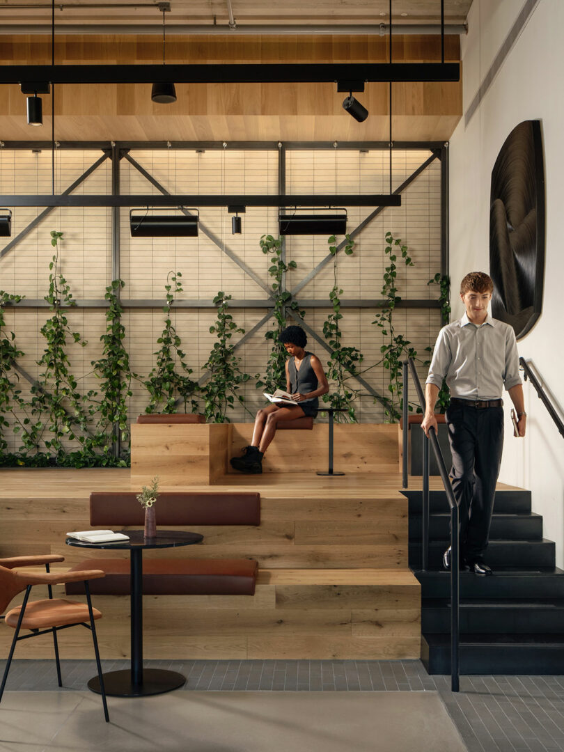A man walks down stairs while another person sits reading on tiered wooden seating in a modern, plant-filled interior space designed by Morris Adjmi Architects.