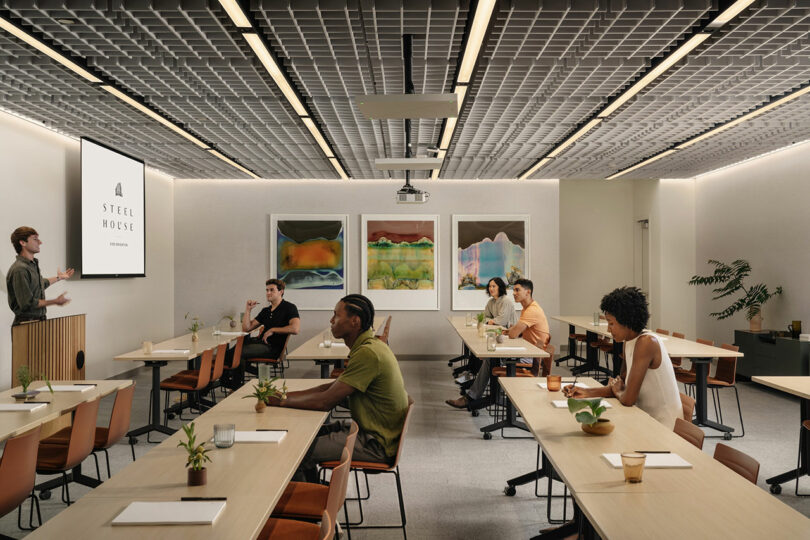 A presenter stands at the front of a modern conference room designed by Morris Adjmi Architects, speaking to six seated attendees. The room features tables, plants, wall art, and a screen displaying 