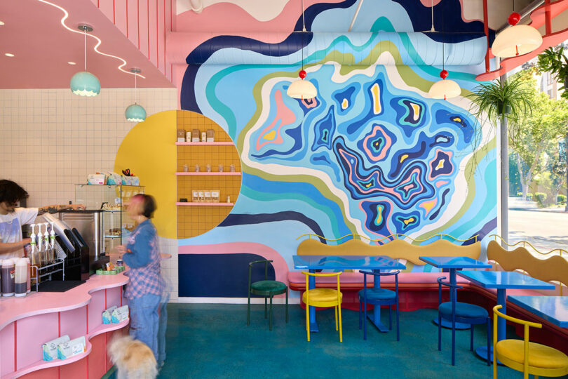 Colorful café interior with abstract mural on the wall, blue tables, yellow and blue chairs, pink counter, hanging lights, and a person ordering at the counter with a dog nearby.