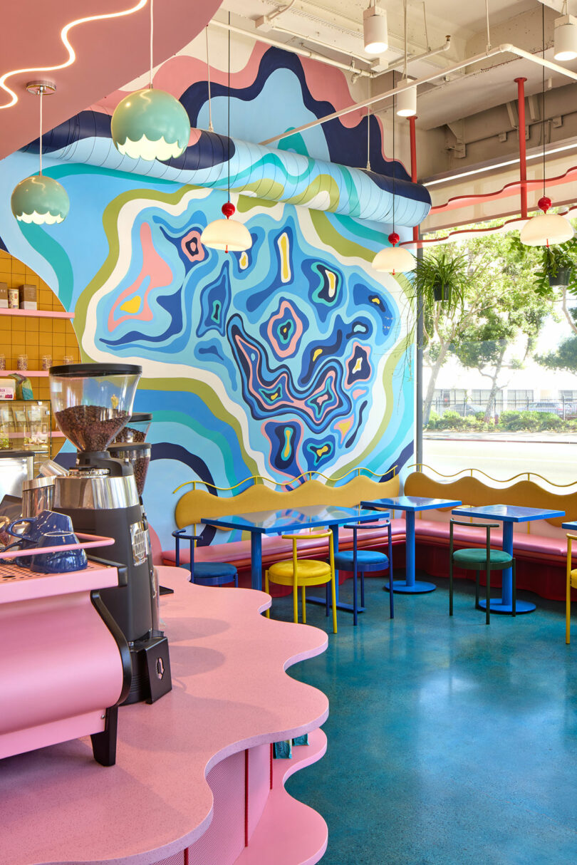 Colorful cafe interior with wavy pink counters, blue floor, multicolored chairs, and a large abstract mural featuring organic shapes in blue, green, and yellow tones on the wall.