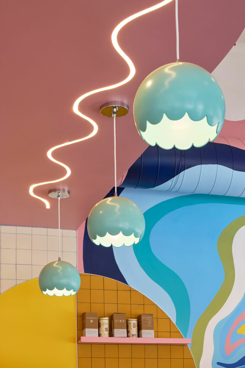 Three spherical pendant lights with scalloped edges hang from a pink ceiling, next to a wavy LED light. Colorful abstract wall art and shelves with boxes are in the background.