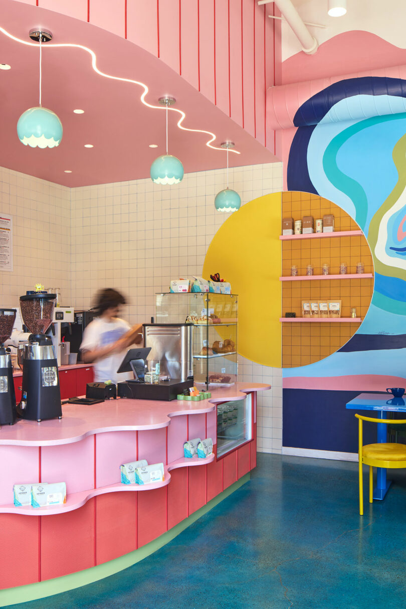 A brightly colored coffee shop interior with a pink counter, blue and yellow wall art, pendant lights, and a staff member preparing drinks behind the counter.