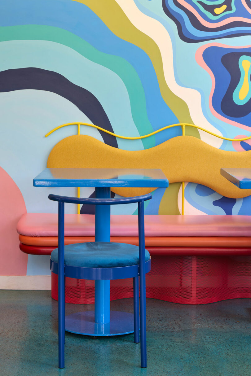 A blue chair and table sit in front of a yellow bench and a colorful, abstract mural with wavy shapes in a modern interior setting.