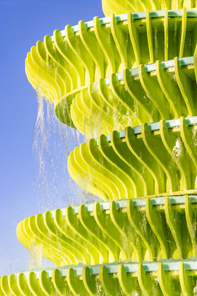 Close-up of a bright yellow-green tiered fountain with water cascading down each level against a clear blue sky.
