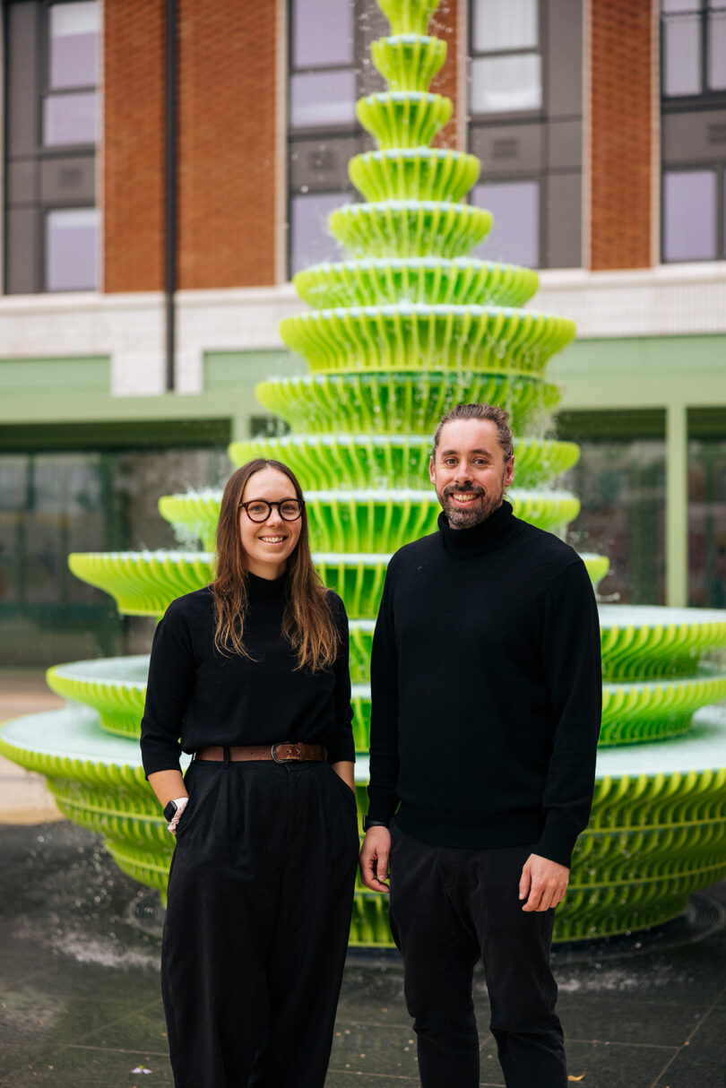 Two people wearing black outfits stand in front of a tiered, bright green water fountain outdoors.