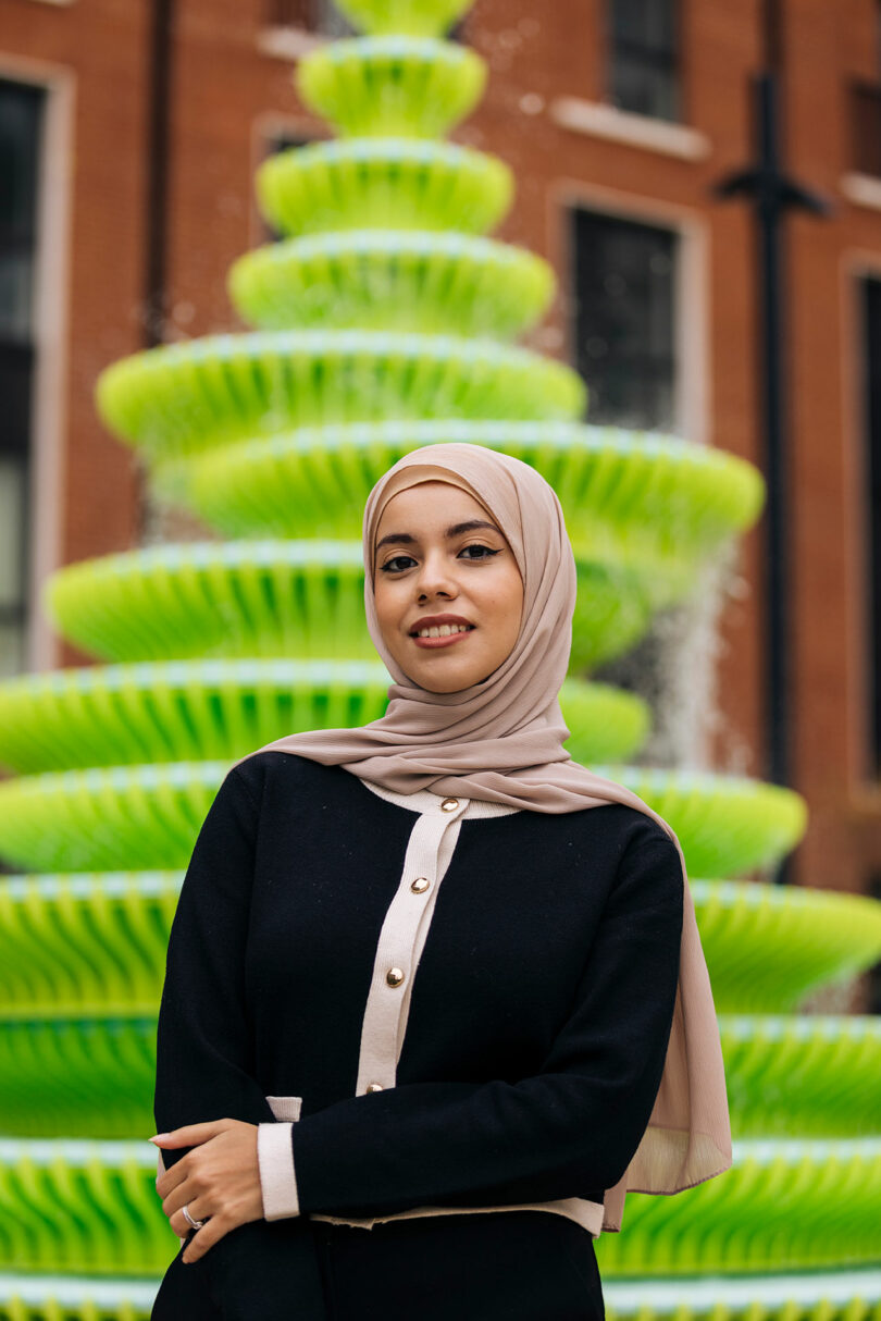 A woman in a beige hijab and black outfit stands in front of a bright green, spiral-shaped outdoor fountain with a brick building in the background.