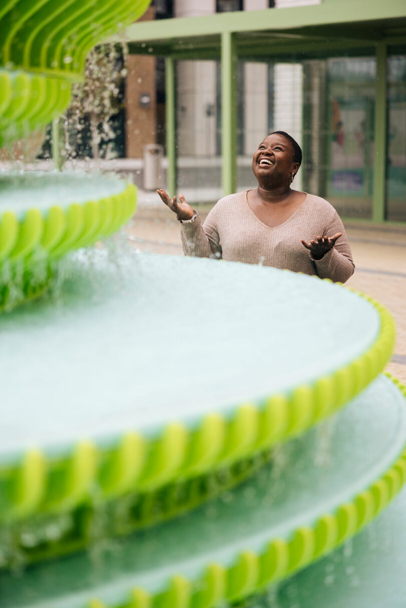 A person stands near a large, tiered green fountain, looking upward with arms raised and a joyful expression.