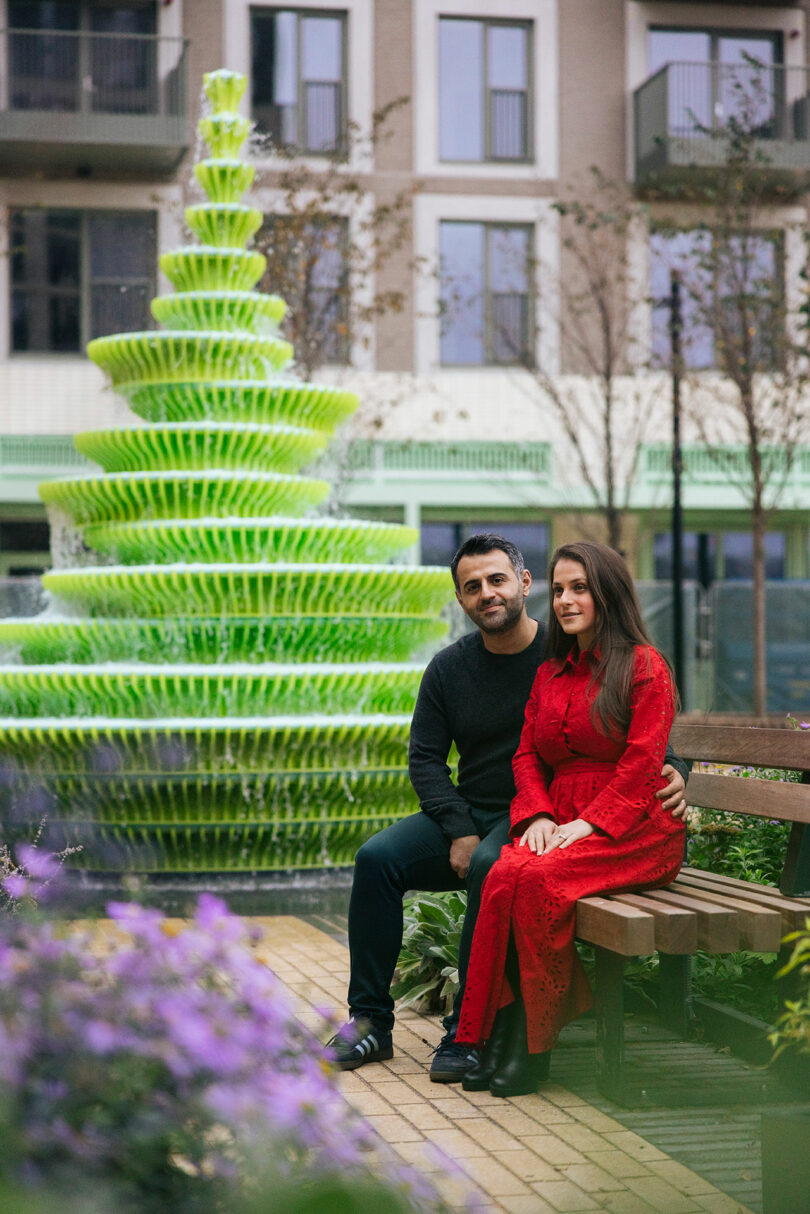 A man and woman sit on a bench near a bright green tiered fountain in an outdoor urban area with buildings and plants in the background.