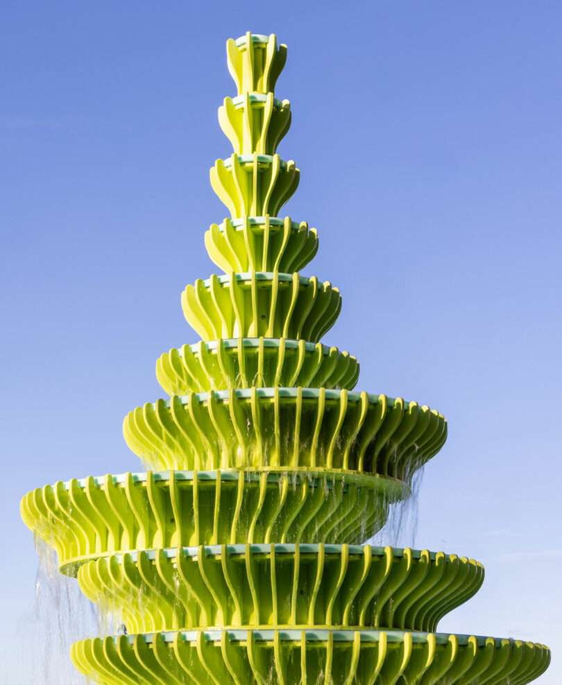 A modern, tiered bright green fountain with cascading water against a clear blue sky.