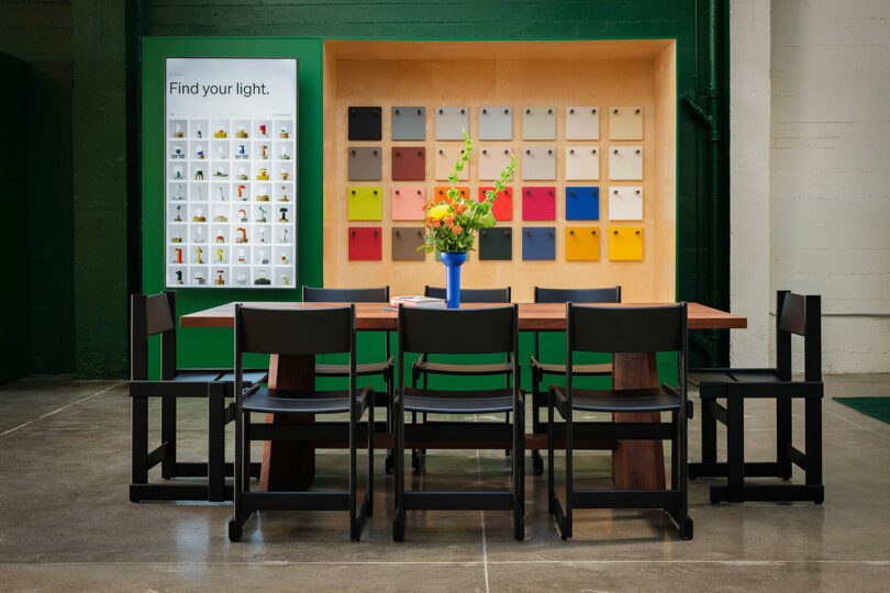A modern meeting room with a wooden table, eight black chairs, a blue vase with flowers, and color sample displays on green and wood-paneled walls.