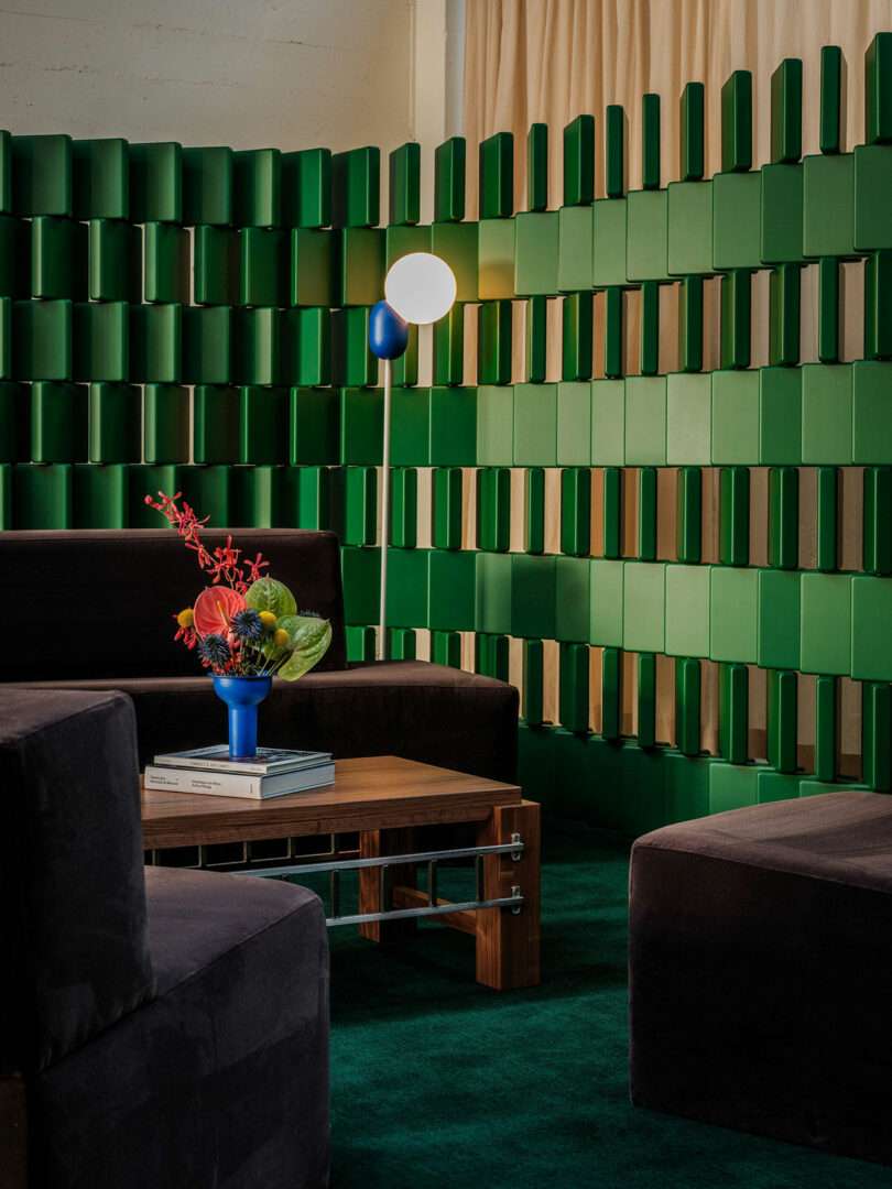 Modern lounge with dark sofas, a wooden coffee table, a blue vase with flowers, books, and a geometric green partition wall; floor lamp with a round white shade in the background.