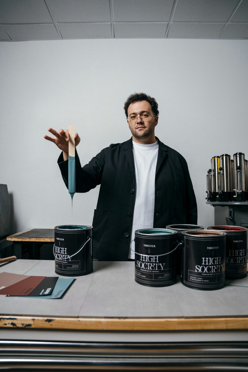 A person in a black coat dips a paint stick into a can labeled 