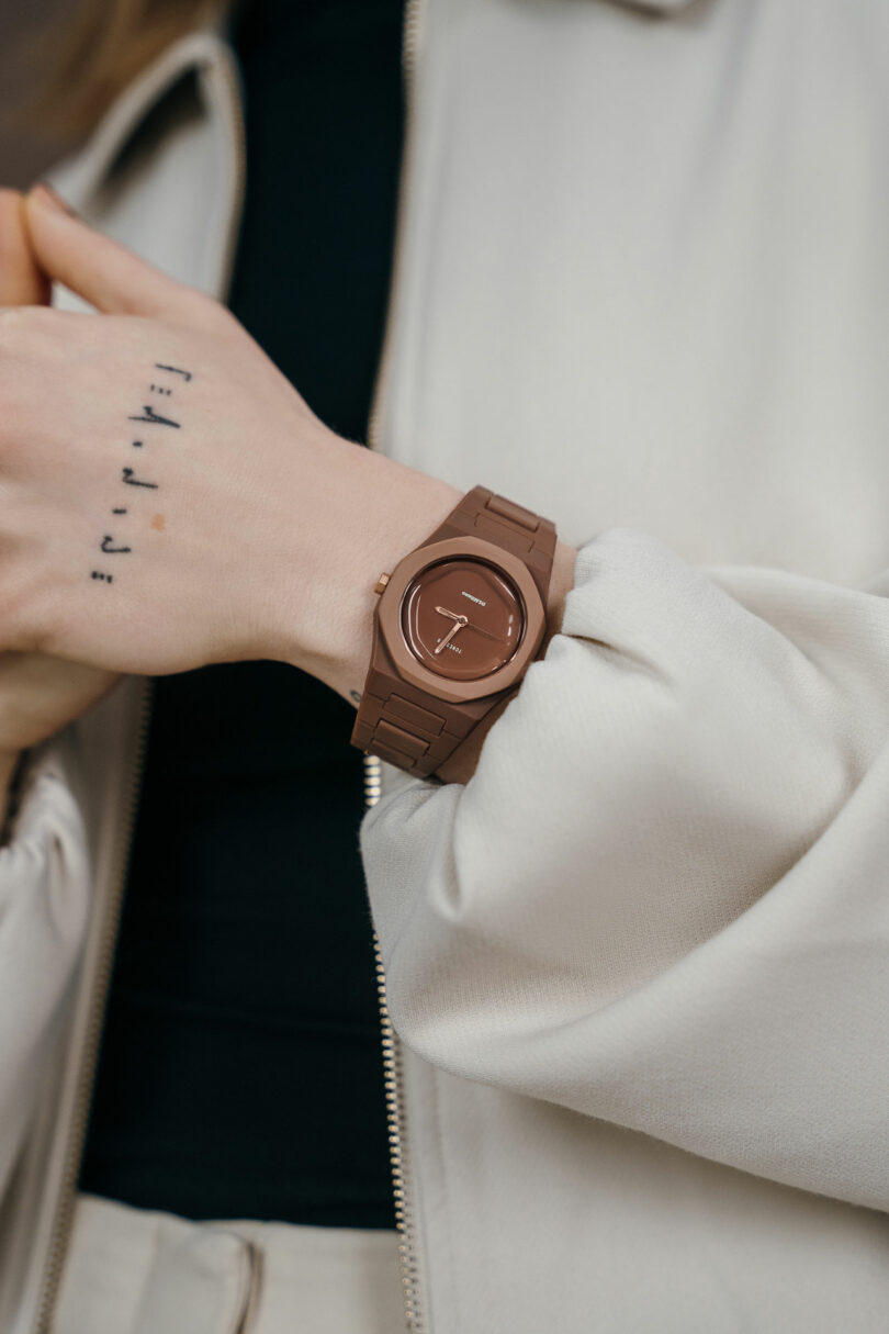 A person wearing a brown wristwatch and light-colored jacket is shown, with small black tattoos visible on their hand.