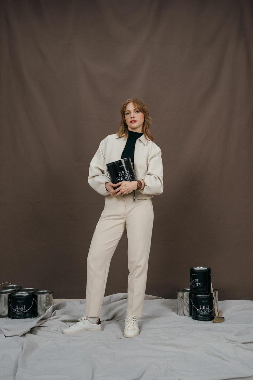 A woman in a cream-colored outfit stands on a draped cloth, holding a black paint can, with additional paint cans and brushes arranged around her against a brown backdrop.