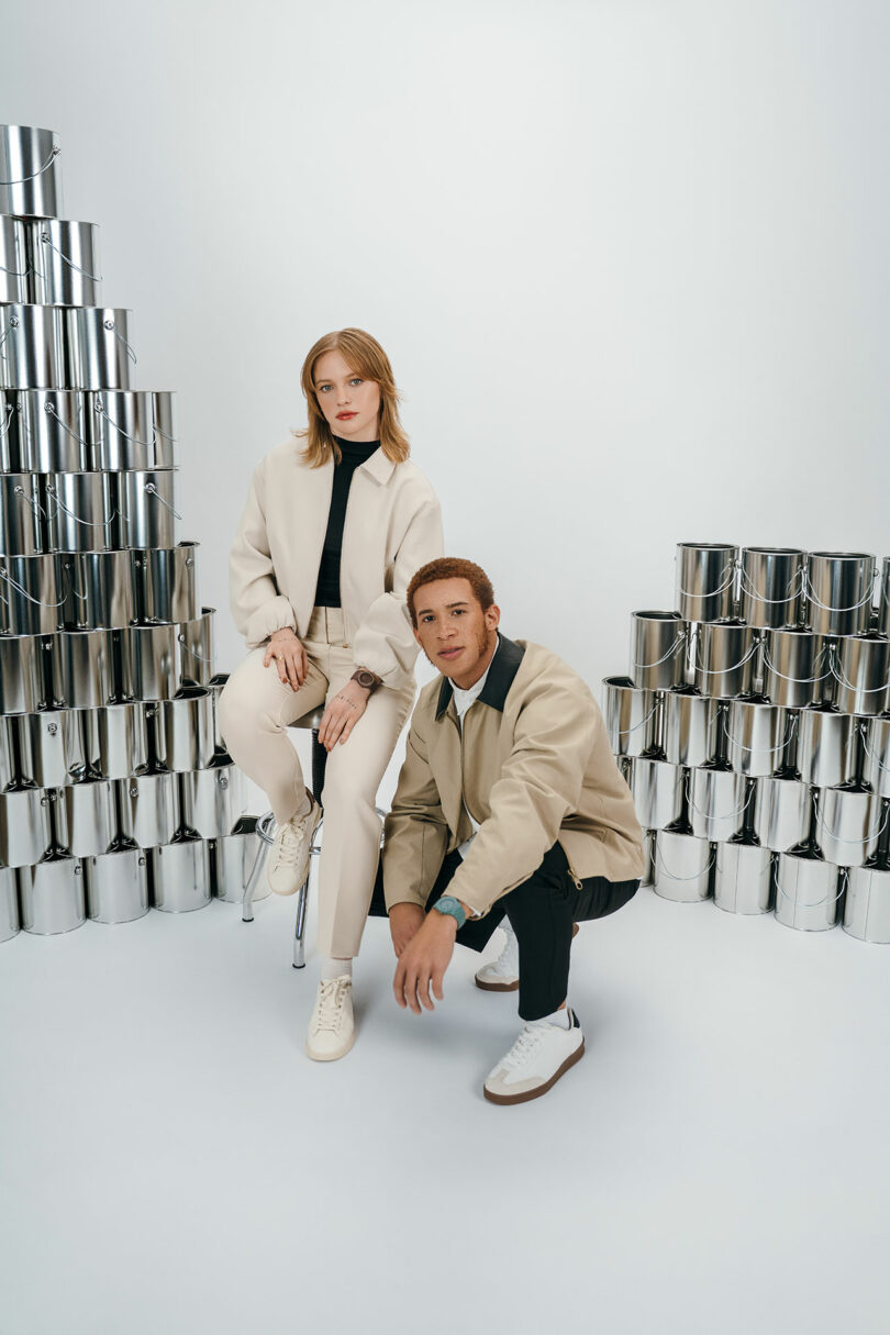 Two people pose in beige outfits; one sits on a stool while the other crouches beside them. They are surrounded by stacked, shiny silver paint cans in a white studio setting.