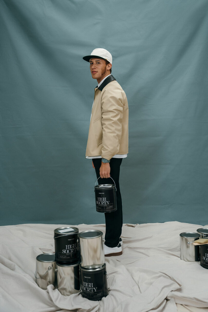 A man in a beige jacket and white cap stands indoors holding a paint can, surrounded by several other paint cans on the floor against a gray backdrop.
