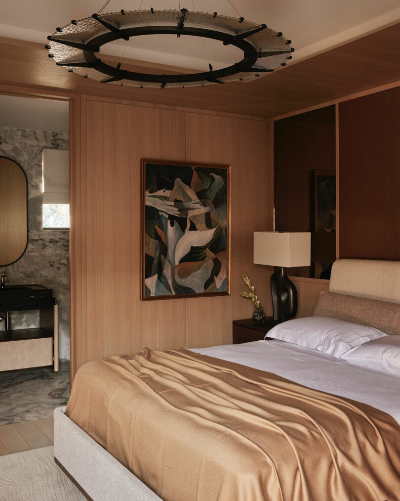 A modern bedroom with wood-paneled walls, an abstract painting, a beige bed with tan bedding, a black lamp, and a glimpse of a marble bathroom in the background.
