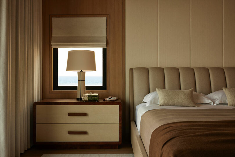 A neatly made bed with tan bedding next to a wooden nightstand with a lamp, a book, and a phone, with a window showing a view of the sea.