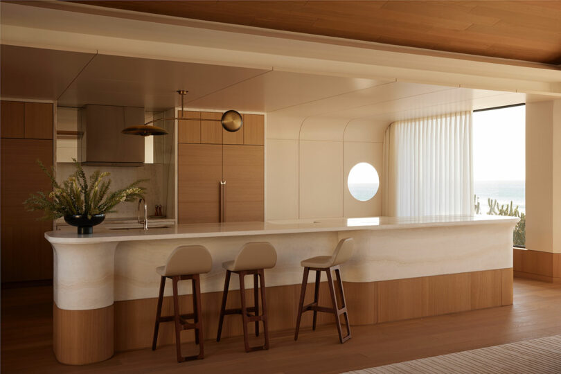 Modern kitchen with light wood cabinetry, a marble island with three barstools, built-in appliances, and a round window letting in natural light.
