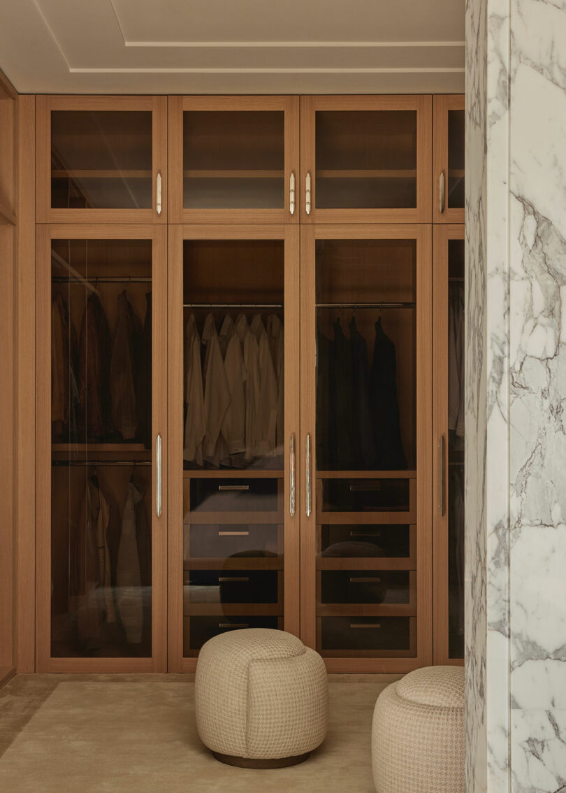 A modern walk-in closet with wooden cabinets, glass doors, hanging clothes, drawers, and two textured round ottomans on a beige carpet. Marble wall partially visible on the right.