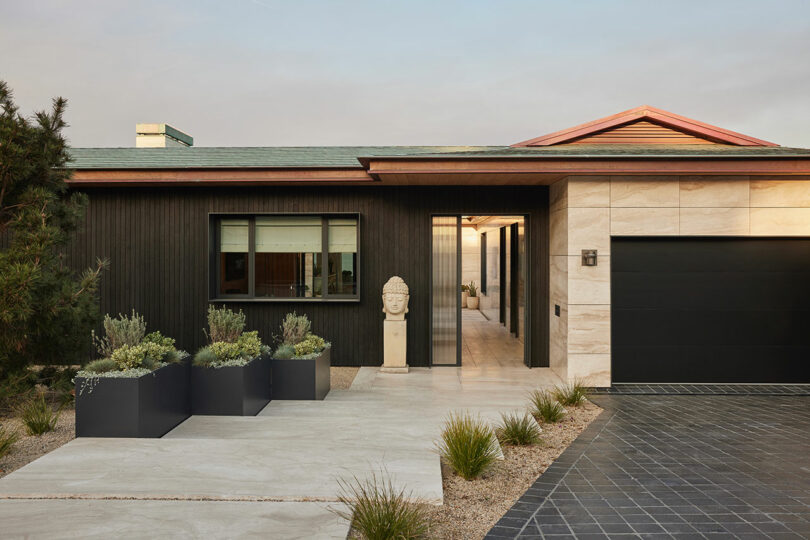 Modern single-story house with black and beige exterior, large window, black garage door, potted plants, and a sculpted bust near the entrance.