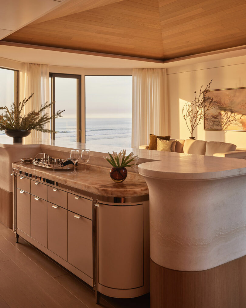 Modern kitchen with marble countertop, built-in cabinets, and decorative plants, featuring large windows with ocean views and warm sunlight filtering into the room.