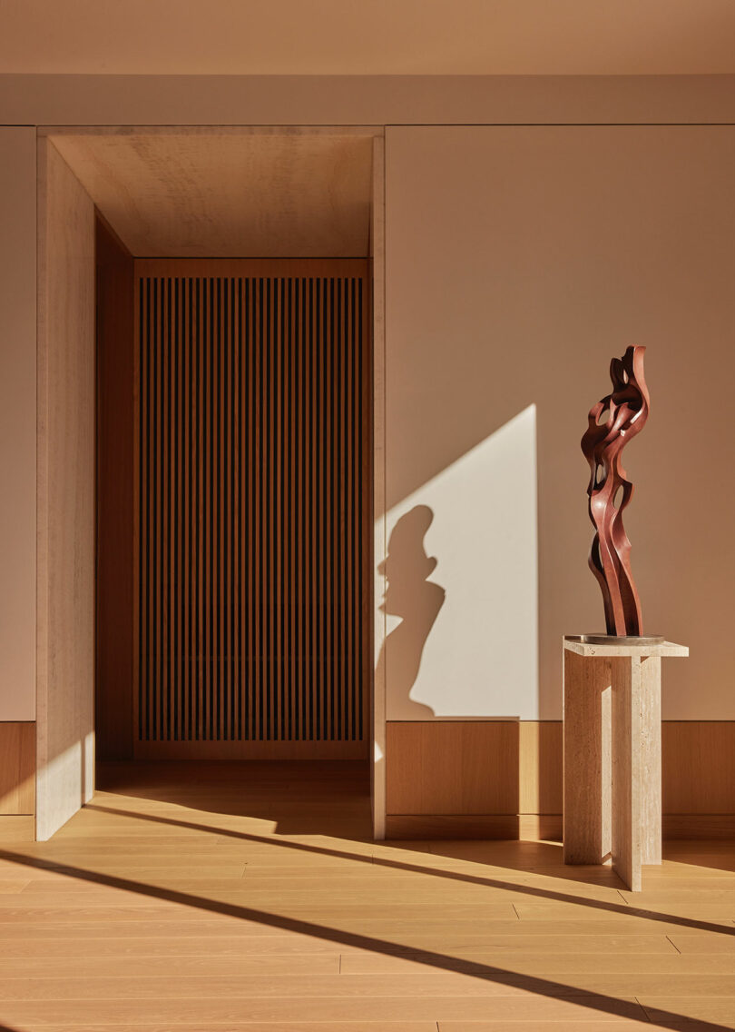 A wooden abstract sculpture on a stone pedestal stands in sunlight near a doorway with vertical wooden slats and beige walls and flooring.
