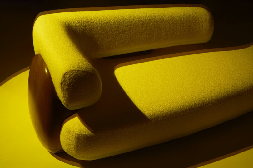 Modern yellow upholstered sofa with rounded edges and a cylindrical wooden armrest, reminiscent of Holly Hunt’s refined aesthetic, photographed under dramatic lighting.