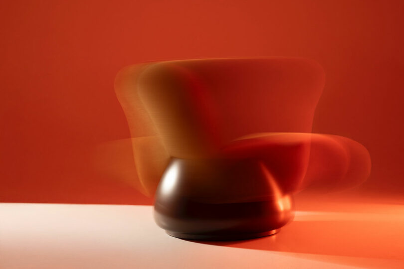 A blurred image of a modern, rounded Holly Hunt chair against an orange background, suggesting motion or rotation, with part of the chair in shadow.