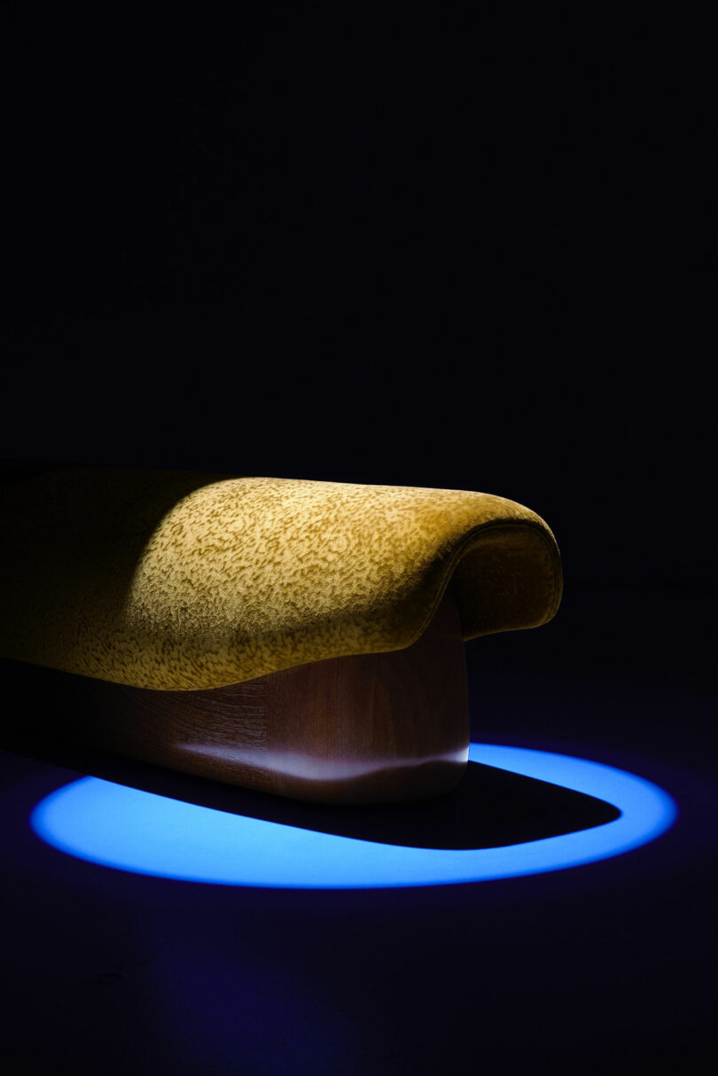A sculptural bench with a yellow speckled cushion and smooth wooden base, illuminated by a blue circular spotlight in a dark setting—echoing the refined aesthetic of Holly Hunt.