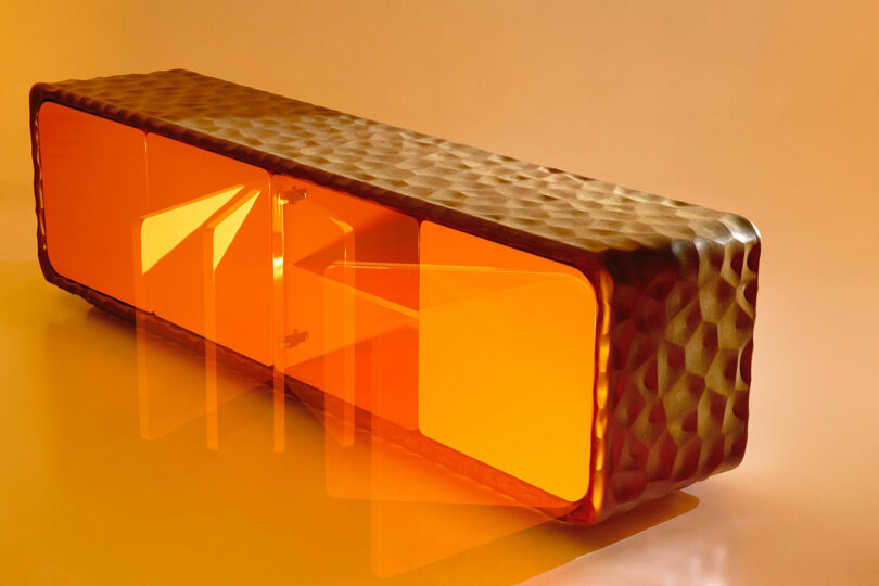 A modern sideboard from Holly Hunt with a textured brown frame and translucent orange doors, two of which are open, set against an orange background.