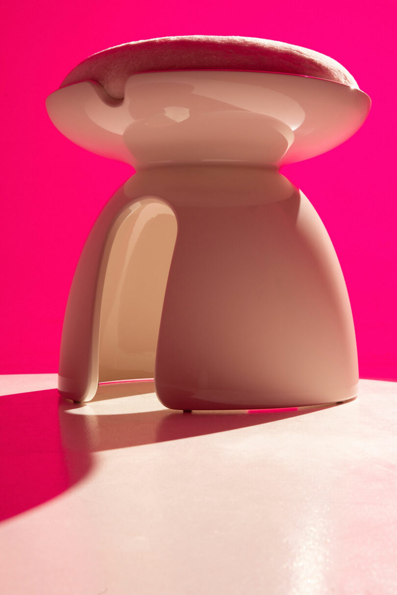 A modern, glossy stool from Holly Hunt with a cutout base and cushioned seat is displayed against a bright pink background.