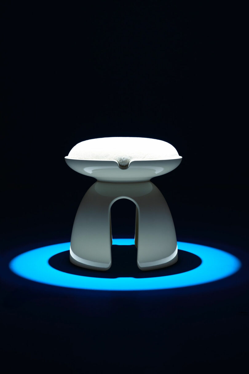 A modern, white, sculptural stool by Holly Hunt is illuminated by a blue spotlight against a dark background.