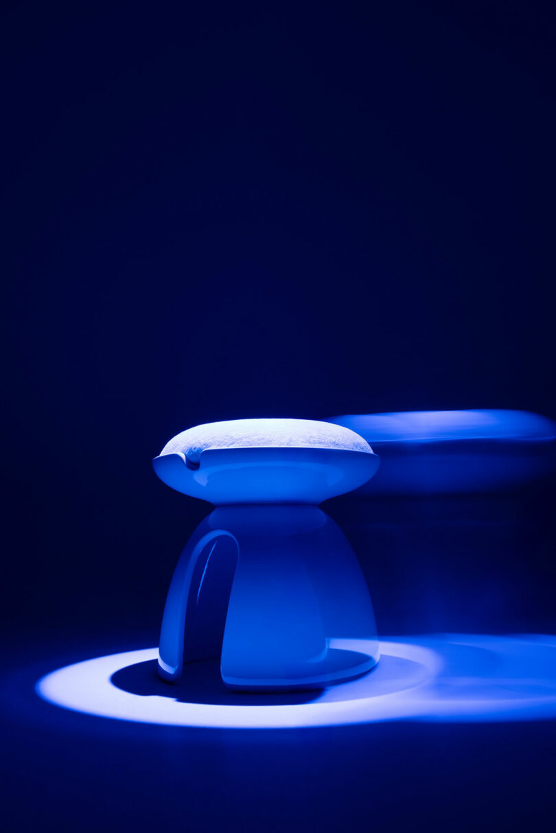 A modern, minimalist, white Holly Hunt stool with a curved design is illuminated by blue lighting against a dark background with a blurred motion effect on one side.