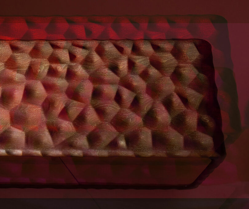 Rectangular object with a textured, geometric surface pattern, illuminated by red and pink lighting. Reminiscent of a Holly Hunt bench or cushion, it is viewed from above at an angle.