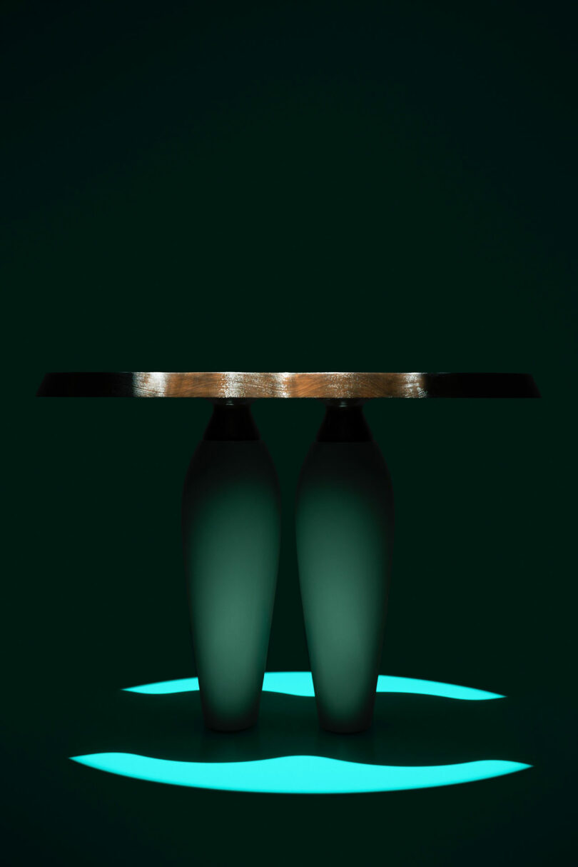 A modern table by Holly Hunt with a thick wooden top and two vase-shaped bases is lit from below, casting teal light and dramatic shadows on a dark background.