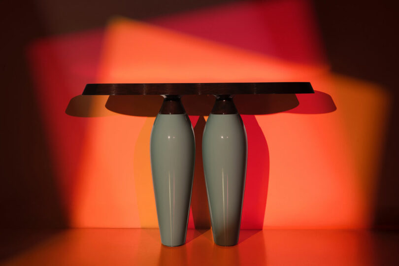 A table with two large, light blue ceramic vase-shaped legs and a dark rectangular top from Holly Hunt is lit with red and orange light, casting colorful shadows on the wall behind.