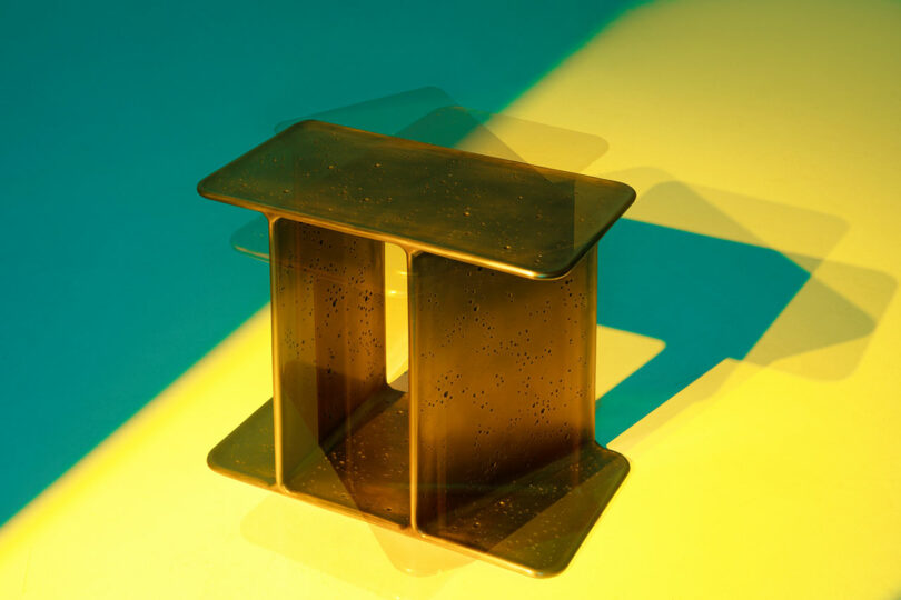 A metallic, rectangular side table by Holly Hunt with rounded edges sits on a yellow surface, casting layered shadows in green and blue hues.