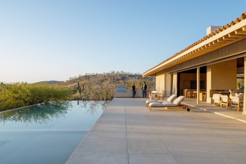 Modern house with an infinity pool and lounge chairs on a spacious patio; two people stand near a railing overlooking a dry, hilly landscape under a clear sky.