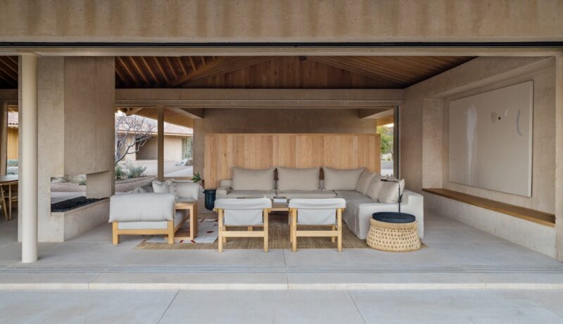 A modern covered patio lounge with light wood and neutral-toned furniture, featuring a sectional sofa, chairs, ottomans, a woven side table, and minimalist décor.