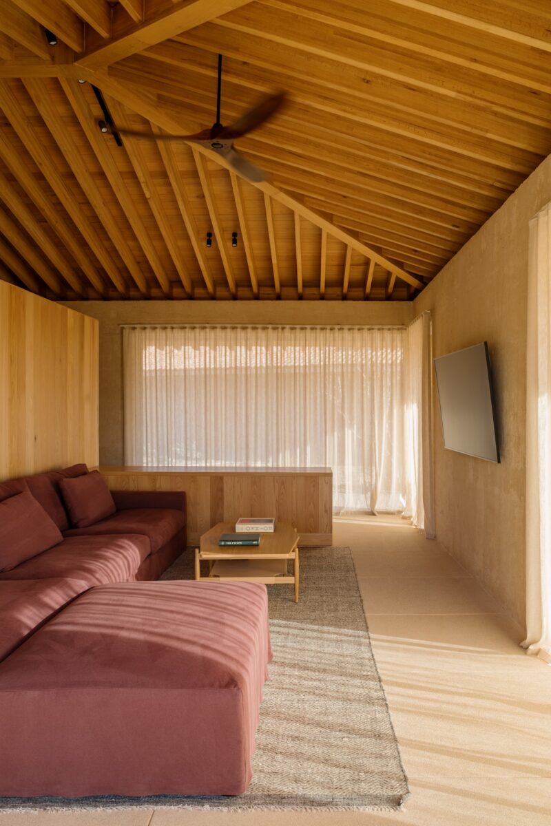 A modern living room with a wooden ceiling, large maroon sectional sofa, coffee table with books, wall-mounted TV, and sheer curtains letting in natural light.