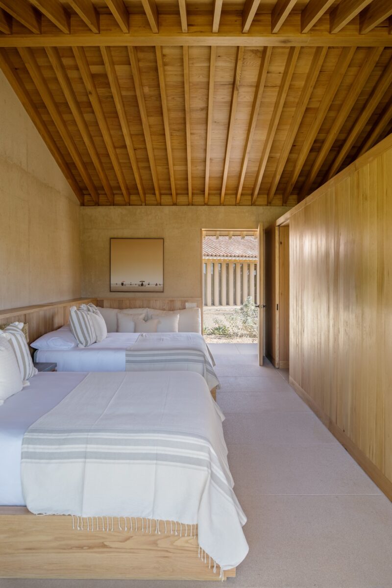 A minimalist bedroom with two beds, striped pillows, wooden walls and ceiling, and a door opening to an outdoor area.