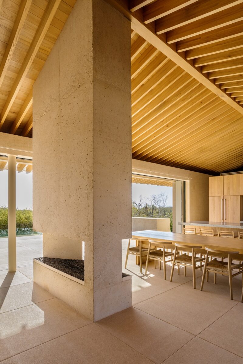 A modern dining area with wooden ceiling, large stone fireplace, wooden table and chairs, and floor-to-ceiling windows overlooking an outdoor landscape.