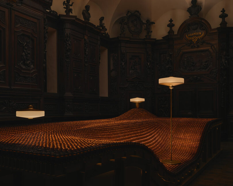A dimly lit room with ornate dark wood paneling features a large, undulating woven surface illuminated by modern floor lamps.