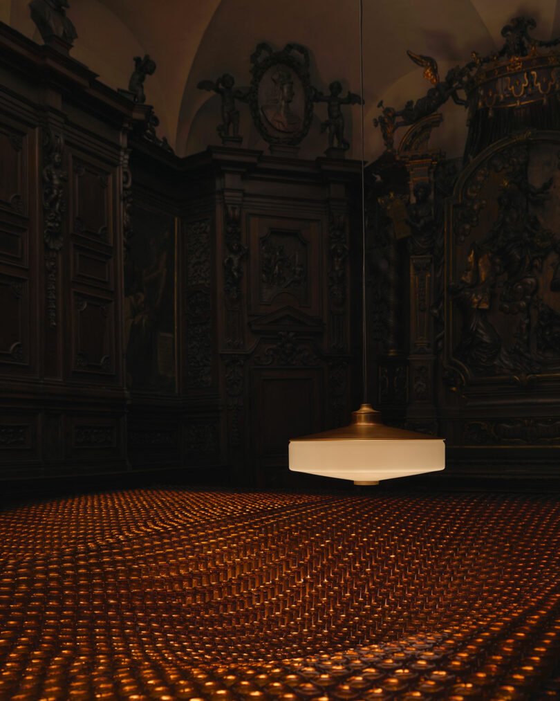 A modern pendant light hangs over a floor covered with a grid of illuminated small bulbs in an ornately decorated, dark wood-paneled room.