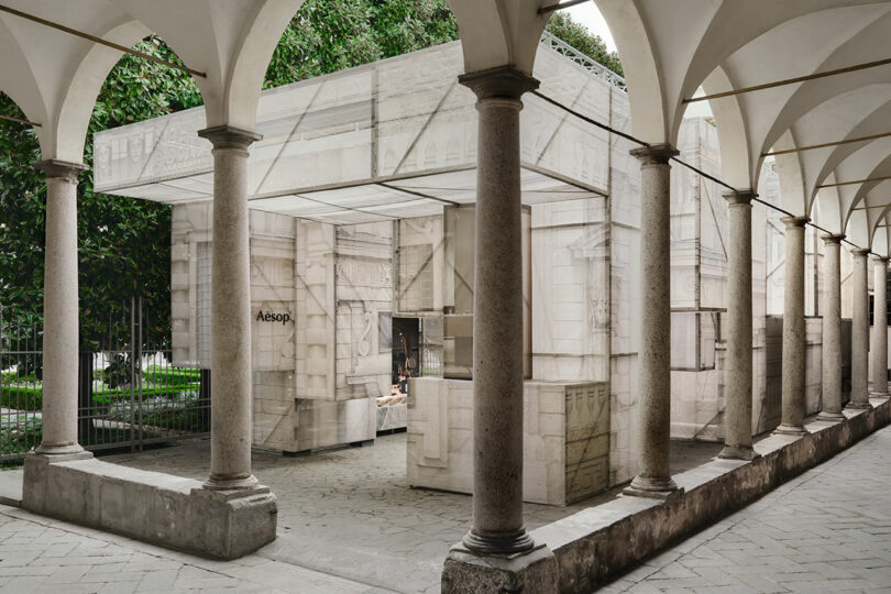 A modern Aesop store with a marble-patterned facade stands beneath arched columns in an outdoor corridor, surrounded by greenery.