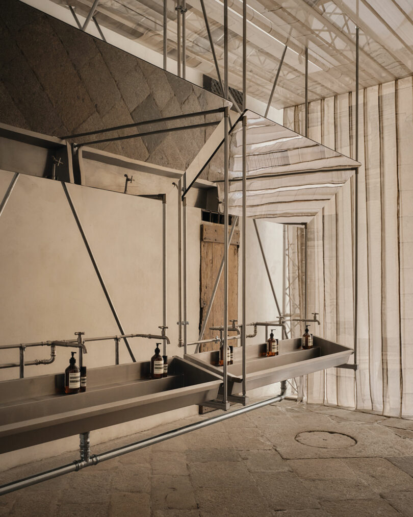 Two industrial-style sinks with metal frames and soap dispensers are installed against a textured wall in a minimalist, open space with natural light.