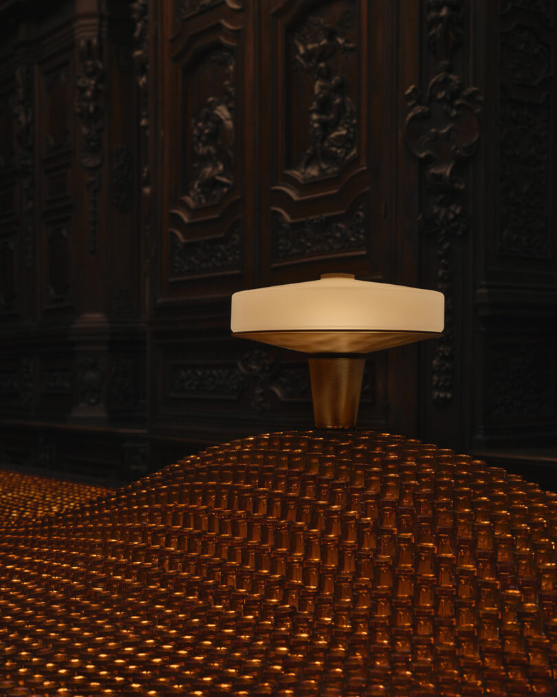 A modern table lamp with a frosted shade sits on a wavy surface of amber glass tiles, with ornate dark wood paneling in the background.